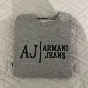 Grå sweatshirt från Armani Jeans - Snygg grå sweatshirt från Armani Jeans med svart logotyp framtill. Tröjan har rund halsringning, långa ärmar och ribbade muddar vid ärmslut och nederkant. Perfekt för en clean och stilren look. Pris kan diskuteras!