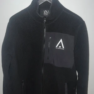 Svart fleecejacka från WearColour XL - Svart fleecejacka från WearColour i storlek XL. Jackan har hög krage, hel dragkedja framtill och en bröstficka med logga och dragkedja. Mjuk och skön fleece med detaljer i syntetmaterial på fickorna. Perfekt för kyliga dagar.