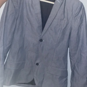 Grå/blå kavaj med två knappar barn - Snygg grå/blå kavaj i barnstorlek 12-13 år, med klassisk passform och två knappar framtill. Kavajen har två fickor med lock och en bröstficka. Perfekt för dig som vill ha en stilren och enkel look. Fodrad insida och diskret design.