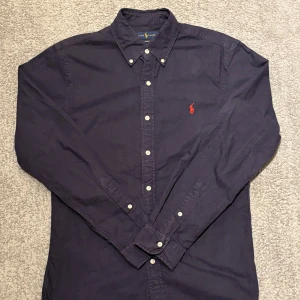 Mörkblå skjorta från Ralph Lauren - Snygg mörkblå skjorta från Ralph Lauren i slim fit med klassisk krage och vita knappar. Ikoniska röda Polo-loggan broderad på bröstet. Skjortan är långärmad och tillverkad i bomull, perfekt för en clean och stilren look.