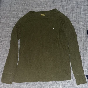 Olivgrön långärmad tröja Polo Ralph Lauren - Säljer en olivgrön långärmad tröja från Polo Ralph Lauren med klassisk vit logga broderad på bröstet. Tröjan har en enkel, stilren design och är tillverkad i mjuk bomull. Perfekt för en avslappnad och snygg look.