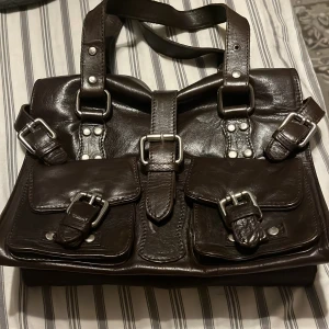 Mörkbrun skinnväska med spännen - Snygg mörkbrun handväska i skinn med flera silvriga spännen och nitar. Väskan har två framfickor med lock och spännen samt dubbla handtag. Perfekt för dig som gillar en cool och robust look med mycket detaljer.