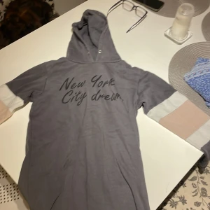 Grå hoodie från Red & Blue med tryck - Grå hoodie från Red & Blue med texten 'New York City dream.' på bröstet. Tröjan har huva och långa ärmar med blockfärgade partier i beige och vitt. Tillverkad i mjuk bomull, perfekt för chill dagar.