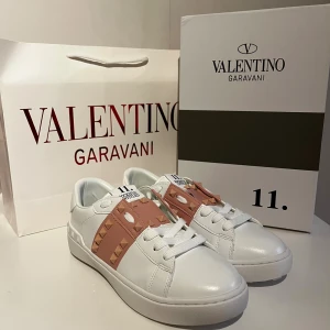 Valentino skor - Snygga Valentino Garavani skor i vitt skinn med breda ljusrosa band och ikoniska nitar. Klassisk rund tå, platt sula och vita snören. Perfekta för dig som vill ha en lyxig och trendig look med extra edge.