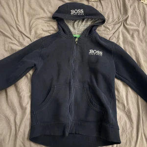Svart hoodie från Hugo Boss - Snygg svart hoodie från Hugo Boss med dragkedja och huva. Tröjan har vita BOSS-loggor på bröstet och huvan, samt två stora fickor framtill. Perfekt för en chill och stilren look.