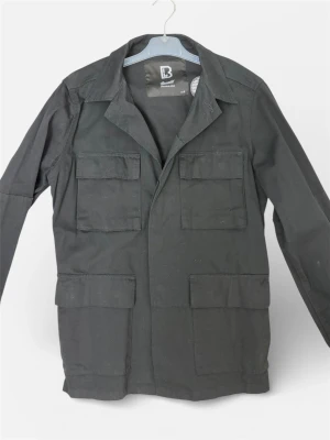 Safarijacka från Brandit - Snygg svart safarijacka från Brandit. Helt och hållet oanvänd och sticker finns kvar i jackan. Stilren och liknar klassiska safarijackorna från Oscar Jacobsson. True to size