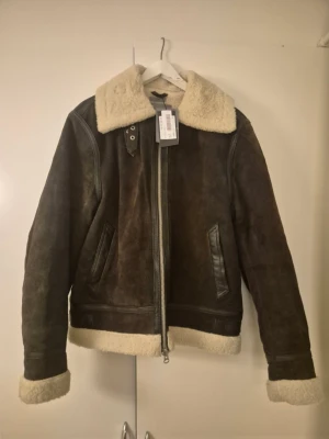 Brun pälsjacka från Bill i mocka - Säljer en brun pälsjacka från Bill i mocka med krage och detaljer i beige fuskpäls. Jackan har dragkedja framtill, två sidofickor med skinnkanter och synliga sömmar. Perfekt för kalla dagar och har en riktigt schysst vintage vibe.