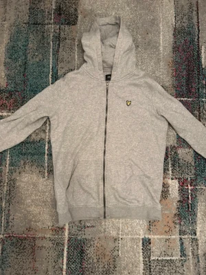 Lyle&Scott kofta - Säljer en klassisk kofta från Lyle & Scott för barn i mycket gott skick. Tillverkad i mjukt och bekvämt material med hög kvalitet som håller formen. Stilren design med den ikoniska örnloggan på bröstet, vilket gör den enkel att matcha både till vardag och mer uppklädda tillfällen.