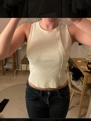 Ribbad vit  linnetopp med knappar på en sida - Säljer en fräsch vit croppad linnetopp med ribbad struktur och dekorativa knappar längs ena axeln. Toppen är ärmlös och har en rund halsringning, perfekt för varma dagar. Materialet känns mjukt och följsamt mot huden.