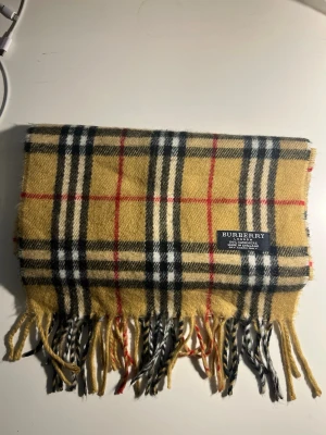 Rutig gul cashmerehalsduk Burberry - Säljer en klassisk gulrutig halsduk från Burberry i 100% kashmir. Den har det ikoniska rutmönstret i svart, vitt och rött samt fransar i ändarna. Perfekt accessoar för att lyfta din outfit med en tidlös brittisk vibe.