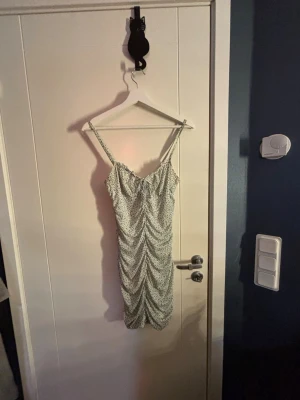 Blommig kortklänning från H&M - Säljer en söt kortklänning från H&M med smala axelband och rynkad front. Klänningen är vit med små blå och gröna blommor, har en figurnära passform och är tillverkad i ett lätt polyestertyg. Perfekt för sommarens alla tillfällen!