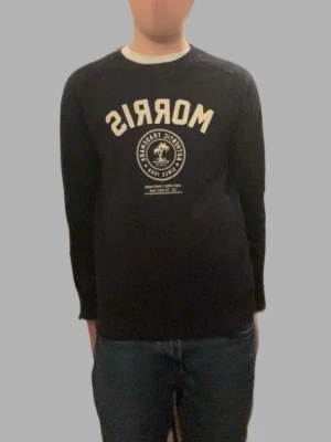 Svart långärmad tröja Morris XS - Svart långärmad tröja från Morris i storlek XS. Tröjan har ett stort vitt tryck med logga och text på bröstet. Rund hals och ribbade muddar vid ärmslut och nederkant. Perfekt för dig som gillar klassisk streetstil och snygga loggor.