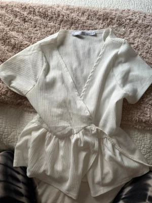 Vit omlottblus från 157 XS - Säljer en vit omlottblus från 157 i storlek XS. Blusen har korta ärmar, ribbad struktur och ett snyggt omlottband i midjan som ger en volangig effekt nedtill. Perfekt för dig som gillar stilrena och feminina detaljer.