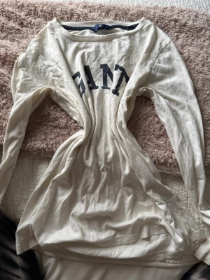 Vit långärmad topp från GANT - Säljer en vit långärmad topp från GANT med svart tryck över bröstet. Toppen har rund halsringning och är tillverkad i mjuk bomull. Perfekt för en chill och stilren look, enkel att matcha med jeans eller kjol.