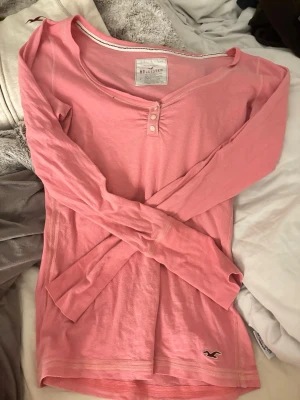 Rosa långärmad topp från Hollister - Säljer en ljusrosa långärmad topp från Hollister med v-ringning och tre små knappar framtill. Toppen har ett broderat Hollister-logo vid nederkanten och är gjord i mjuk bomull med lite stretch. Perfekt för chill dagar och enkel att matcha med jeans.