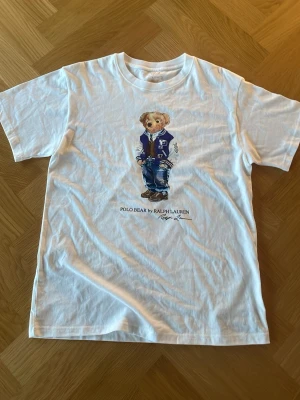 Vit Polo Bear t-shirt från Ralph Lauren - Vit t-shirt från Ralph Lauren med ikoniskt Polo Bear-tryck på bröstet. Klassisk passform och rund hals. Tillverkad i mjuk bomull som känns skön mot huden. Perfekt för dig som gillar streetstyle och vill ha en stilren men lekfull look.