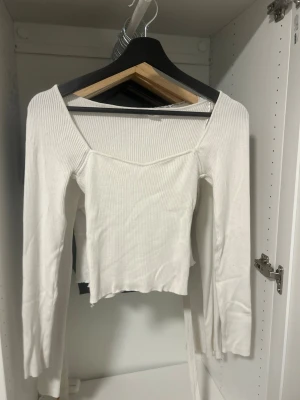 Vit ribbad långärmad topp - Säljer en vit ribbad långärmad topp med v-ringad hals och tight passform. Materialet är mjukt och stretchigt, perfekt för dig som gillar en enkel men snygg stil. Toppen har en modern look med sin ribbade struktur och lite kortare längd. Endast använd en gång 