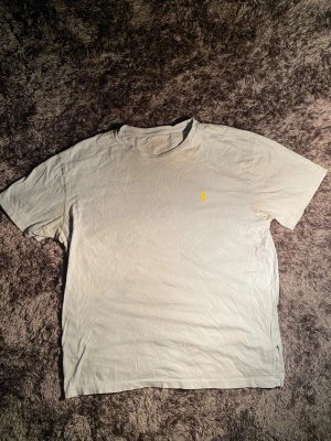 Vit t-shirt från Polo Ralph Lauren - Vit t-shirt från Polo Ralph Lauren i 100% bomull. Klassisk rund hals och korta ärmar, med gul broderad logga på bröstet. Enkel och clean design, perfekt till jeans eller shorts. Märkesetikett i sidan.