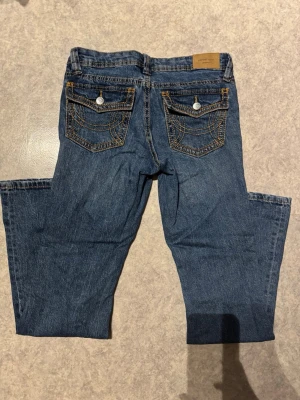 Blå jeans från Perfect Jeans, storlek 34 - Säljer ett par klassiska blå jeans från Perfect Jeans i storlek 34. Jeansen har snygga bakfickor med dekorativa sömmar och knappar, samt femficksdesign och kontrastfärgade stickningar. Modellen har rak passform och är tillverkad i slitstarkt denimtyg. Mått: 	•	Midjemått: ca 37 cm 	•	Grenhöjd: ca 18 cm 	•	Innerbenslängd: ca 76 cm