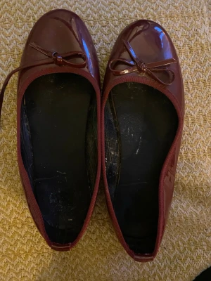 Vinröda ballerinaskor med rosett - Säljer ett par vinröda ballerinaskor med glansig finish och rund tå. Skorna har en söt rosettdetalj framtill och platt sula. Materialet är syntet och insidan är svart. Perfekt för dig som gillar klassisk och enkel stil.