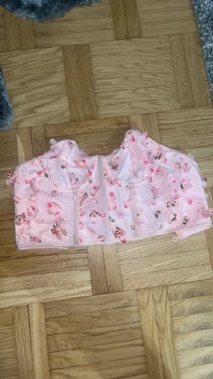 Rosa blommig korsett med snörning - Säljer en ljusrosa korsett med 3D-blommor i rosa och beige nyanser. Toppen har snörning i ryggen och är croppad. Materialet är syntet med genomskinliga partier och dekorativa blommor som ger en romantisk vibe. Perfekt för dig som vill sticka ut med en söt och unik topp.