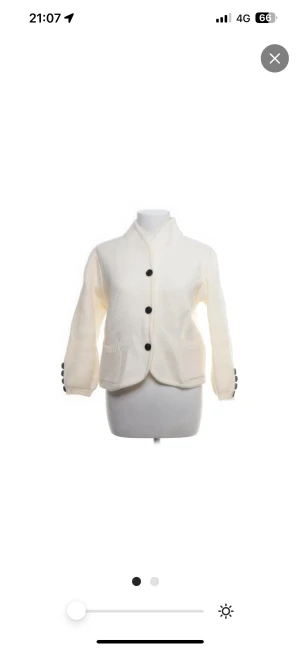 Vit cardigan jacka med svarta knappar - Säljer en vit cardigan jacka med snygga svarta knappar framtill och på ärmsluten. Jackan har två fickor och en cool krage som ger en chill vibe. Materialet är stickat och känns mjukt och skönt. Passformen är normal och ärmarna är långärmade.