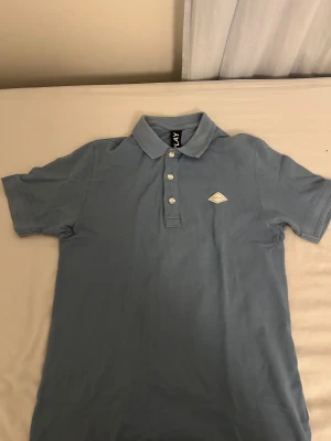 Blå piké från Replay - Säljer en stilren blå pikétröja från Replay med klassisk krage och tre knappar framtill. Tröjan har korta ärmar och ett snyggt Replay-märke på bröstet. Materialet är mjukt och känns som bomull, perfekt för en chill look.