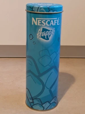 Nescafé frappé metallburk  - En blå Nescafé frappé burk. Burken är av metall och den är i jätte bra skick. Nescafé frappé metallburken kan användas som dekoration och som förvaring.