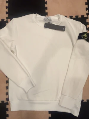 Vit sweatshirt från Stone Island - Säljer en clean vit sweatshirt från Stone Island med klassisk patch på ärmen. Tröjan har rund hals, långa ärmar och ribbade muddar. Perfekt för dig som gillar stilrena plagg med street-vibe och ikonisk logga.