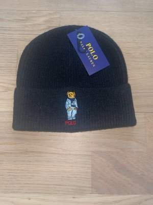 Svart stickad mössa från Polo Ralph Lauren - Svart ribbstickad mössa från Polo Ralph Lauren med broderad nalle på framsidan och uppvikt kant. Märkeslapp på insidan. Perfekt för kalla dagar och snygg detalj till din outfit.