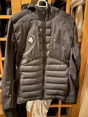 Svart pufferjacka från Sail Racing - Svart pufferjacka från Sail Racing med huva och snygga logodetaljer på bröstet och ryggen. Jackan har quiltad framsida, dragkedja och patch med Antarktis-expedition. Perfekt för kalla dagar och har en modern, sportig look.   Oanvänd ny skick!