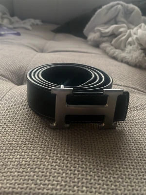 Svart skärp med H logotyp - Stilrent svart skärp med ett stort H-format metallspänne. Skärpet har vita sömmar längs kanterna och är tillverkat i slätt skinn. Perfekt accessoar för att lyfta din outfit med en lyxig touch.