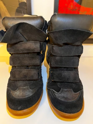 Svarta sneakers från Isabel Marant - Säljer ett par svarta höga sneakers från Isabel Marant med cool design och bruna gummisulor. Skorna har flera breda kardborreband, rund tå och är tillverkade i mocka och skinn. Insidan är mjukt fodrad i ljust skinn. Perfekt för dig som gillar statement-skor.