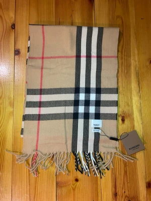 Beige rutig Burberry scarf i kashmir - Säljer en klassisk beige halsduk från Burberry med det ikoniska rutiga mönstret i svart, vitt och rött. Halsduken är tillverkad i 100% kashmir och har fransar längs nederkanten. Perfekt för dig som gillar lyxiga och tidlösa plagg med snygg design.