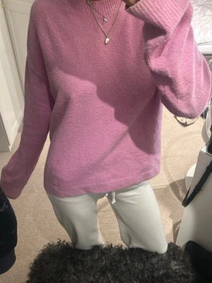 Ljusrosa stickad tröja med rund hals - Mysig ljusrosa stickad tröja med rund hals och långa ärmar. Tröjan har en loose passform och ribbade muddar vid hals, ärmslut och nederkant. Perfekt för att hålla sig varm under kyliga dagar.