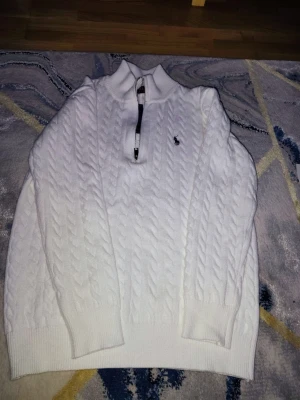 Vit stickad tröja från Ralph Lauren - Säljer en vit stickad tröja från Ralph Lauren med klassiskt kabelmönster och half zip framtill. Tröjan har långärmad design, ribbade muddar och liten broderad logga på bröstet. Perfekt för dig som gillar stilrena och tidlösa plagg.