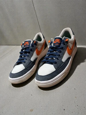 Nike Adversary SB Premium - Nike Adversary SB Premium sneakers med grå och vita paneler i mocka och bomull, samt orange swoosh och detaljer. Lågt skaft, marinblå snörning och rund tå. Snygg kontrast mellan färgerna och klassisk Nike SB logga på plösen. Perfekt för dig som gillar streetwear. Skorna är använda men i bra skick. Nypris 1000kr