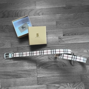 Rutig Burberry bälte i beige och svart - Säljer ett klassiskt Burberry bälte med det ikoniska rutiga mönstret i beige, svart, vitt och vinrött. Bältet har en silverfärgad metallspänne med Burberry-logga. Kommer i original ask med certifikat.