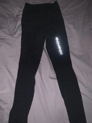 Svarta leggings från Ninenine - Säljer ett par svarta leggings från Ninenine med reflekterande logga på benet. De har hög midja och är gjorda i ett stretchigt syntetmaterial som passar perfekt för träning. Snygg och enkel design med tight passform.