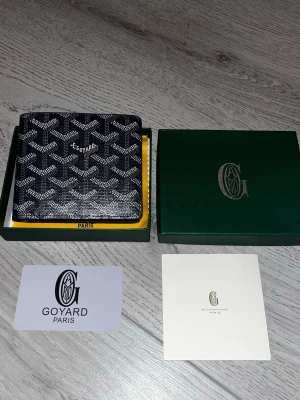 Svart G plånbok med mönster - Säljer en stilren mörkblå plånbok från G med ikoniskt grafiskt mönster i vitt och grått. Plånboken har flera kortfack och är tillverkad i exklusivt skinn. Kommer med original ask, tygpåse och äkthetskort. Perfekt för dig som gillar lyxiga accessoarer.