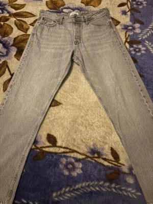 Grå jeans från Jack & Jones - Säljer ett par grå jeans från Jack & Jones med klassisk femficksdesign och bälteshällor. Jeansen har rak passform och är tillverkade i slitstarkt denim. Snygga detaljer på bakfickorna och en neutral grå ton som passar till det mesta.