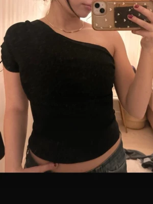 Svart oneshoulder topp - Säljer en svart oneshoulder topp med tight passform. Toppen har en kort ärm på ena sidan och är axelbandslös på andra sidan, vilket ger en cool asymmetrisk look. Perfekt att styla med jeans eller kjol för en trendig outfit.