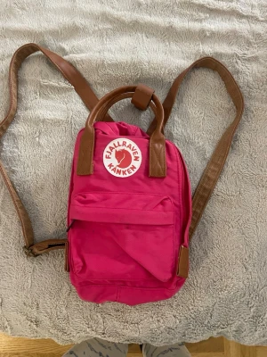 Rosa Kånken ryggsäck från Fjällräven - Säljer en snygg rosa Kånken ryggsäck från Fjällräven med bruna detaljer och axelremmar. Väskan har ett klassiskt rundat handtag, framficka med lock och det ikoniska Fjällräven-märket framtill. Perfekt storlek för vardagsgrejer och enkel att bära.