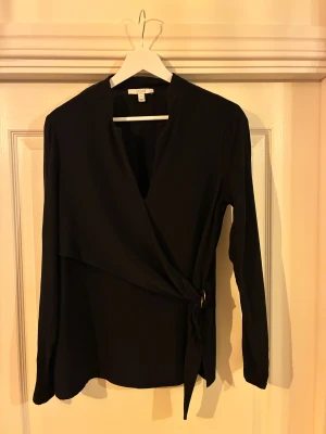 Exklusiv svart blus från House Of Dagmar - Exklusiv och fin svart blus från House Of Dagmar i 100% Viscose, mjukt, glansigt material. Långärmad modell med knyt i sidan och en liten metallring som detalj. Perfekt för dig som gillar stilrena plagg med en twist. Perfekt för utgång eller finmiddag. Nypris 3400 kronor Använd fåtal gånger, inga tecken på slitage eller användning