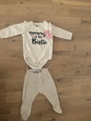 Sött set med body och byxor Vit babyset med text och rosett - Ett sött set bestående av en body med lång ärm och texten ”Mommy is my Bestie” samt ett par matchande byxor. Bodyn har en liten volangdetalj vid axlarna och en dekorativ rosett. Supersött babyset med vit långärmad body från Miniworld med texten 'mommy is my bestie' och rosa prickiga rosetter samt små rosa hjärtan. Tillhörande vita byxor från Dirkje med resår i midjan. Perfekt för små fashionistas!