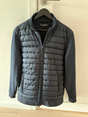 Navy blå cardigan från Dressmann - Navy blå jacka från Dressmann med quiltad framsida och släta ärmar. Jackan har dragkedja hela vägen, ståkrage och två sidofickor. Materialet är polyester och jackan har en clean, sportig look som funkar till många stilar.