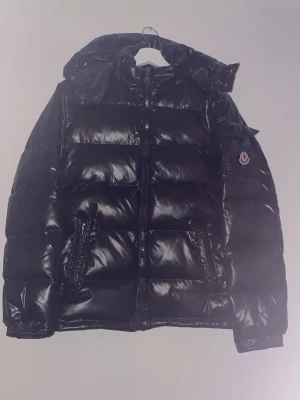 Svart Moncler pufferjacka med huva - Säljer en svart pufferjacka från Moncler med glansig finish och huva. Jackan har dragkedja framtill, två fickor med dragkedja och Moncler-logga på ärmen. Perfekt för kalla dagar och riktigt snygg till streetwear-stilen.