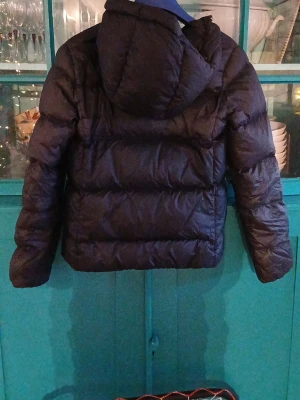 Svart pufferjacka från GANT - Svart pufferjacka från GANT med huva och två fickor framtill med knappstängning. Jackan har quiltad design och dragkedja. Perfekt för kalla dagar och har en schysst, lite loose passform.