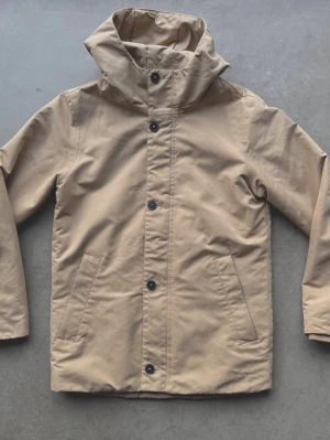 Elvine herrjacka S - Beige vinterjacka i kortare parkasmodell från Elvine i storlek herr S. Jackan har huva, dragkedja och knappar framtill samt två snedställda fickor. Vind- och vattentät med tejpade sömmar på utsatta ställen. Tillverkad i polyester med Thermore-foder för extra värme.  Diskret fläck som inte går bort strax nedanför på ena armbågen, men inget som stör, se bild.