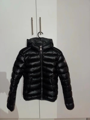Svart Colmar pufferjacka med huva - Säljer en svart pufferjacka från Colmar i storlek 40. Jackan har en glansig finish, huva och dragkedja framtill. Märket syns tydligt på ärmen och jackan är fylld med naturligt dun. Perfekt för kalla dagar och har en riktigt snygg streetstil.